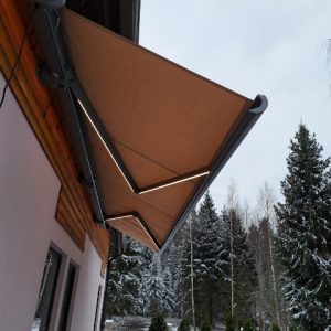 Ruskea retractable-markiisi taloverannalla talvisessa maisemassa, taustalla lumisia kuusia