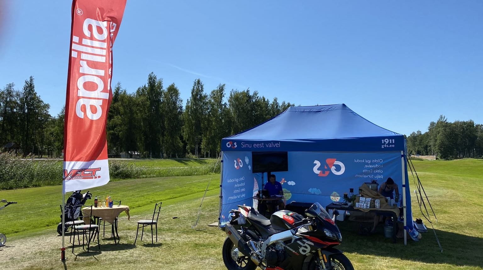 Moottoripyörä näyttelyteltalla kesäpäivällä, punainen Aprilia-lippu ja sininen G&S teltta vihreällä