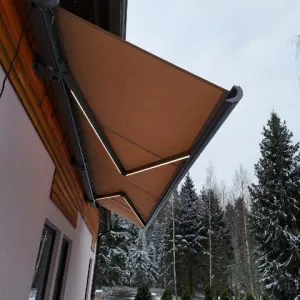 Ruskea retractable-markiisi taloverannalla talvisessa maisemassa, taustalla lumisia kuusia